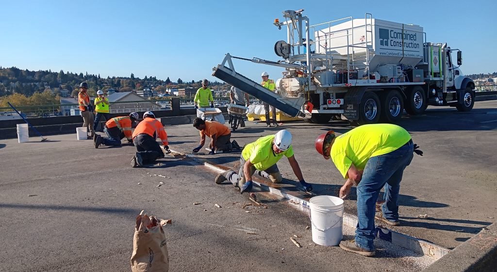 Several construction workers wearing yellow and orange personal protective equipment work on a part of a bridge on a bright sunny day. A large construction vehicle is in the background.
